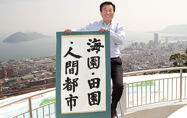 高松市街、瀬戸内海を背に自身の書を掲げる大西市長＝高松市西宝町の峰山公園展望台